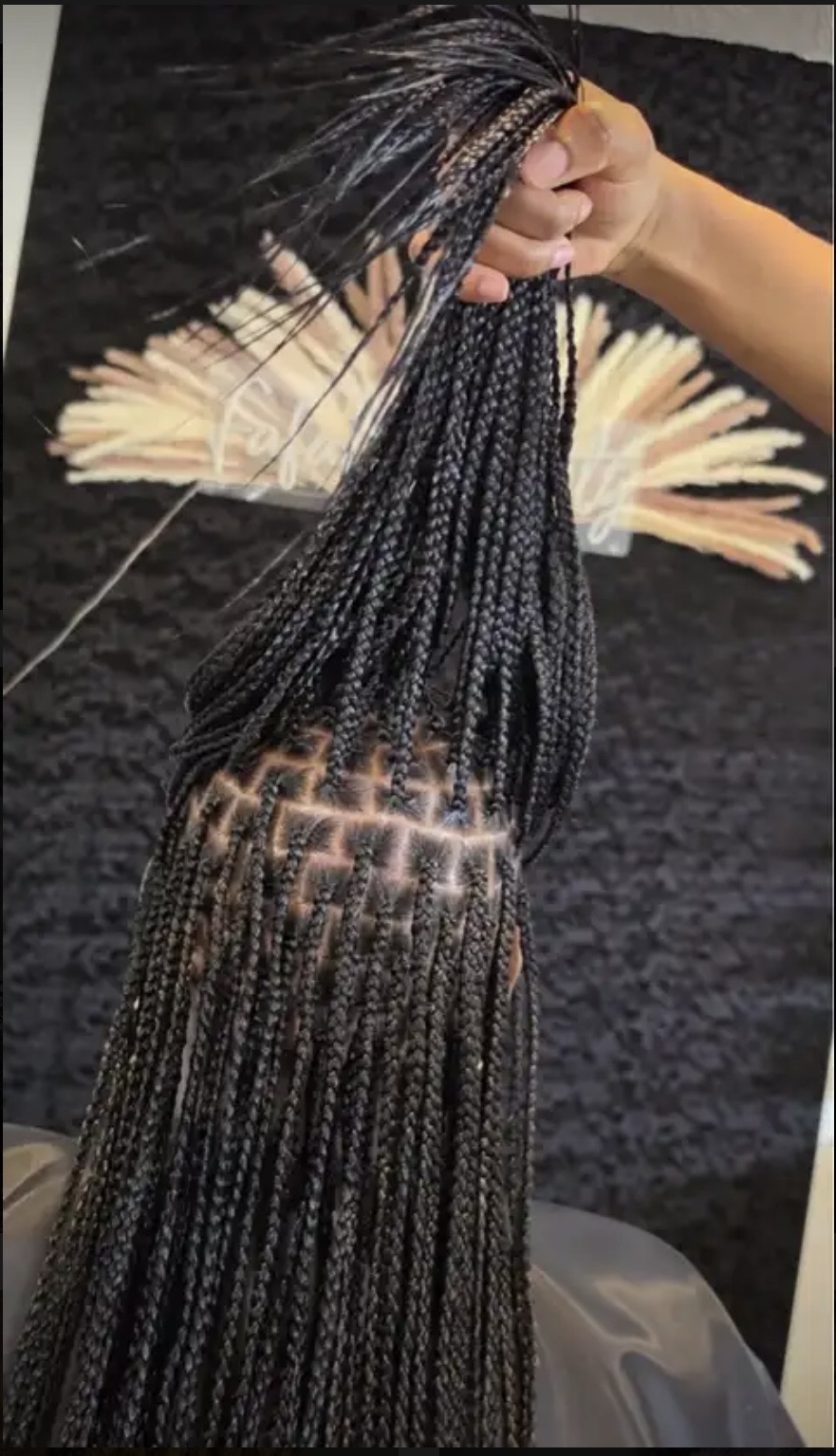 Person holding braided hair with a patterned bag, black textured background, decorative fan, and black wall.