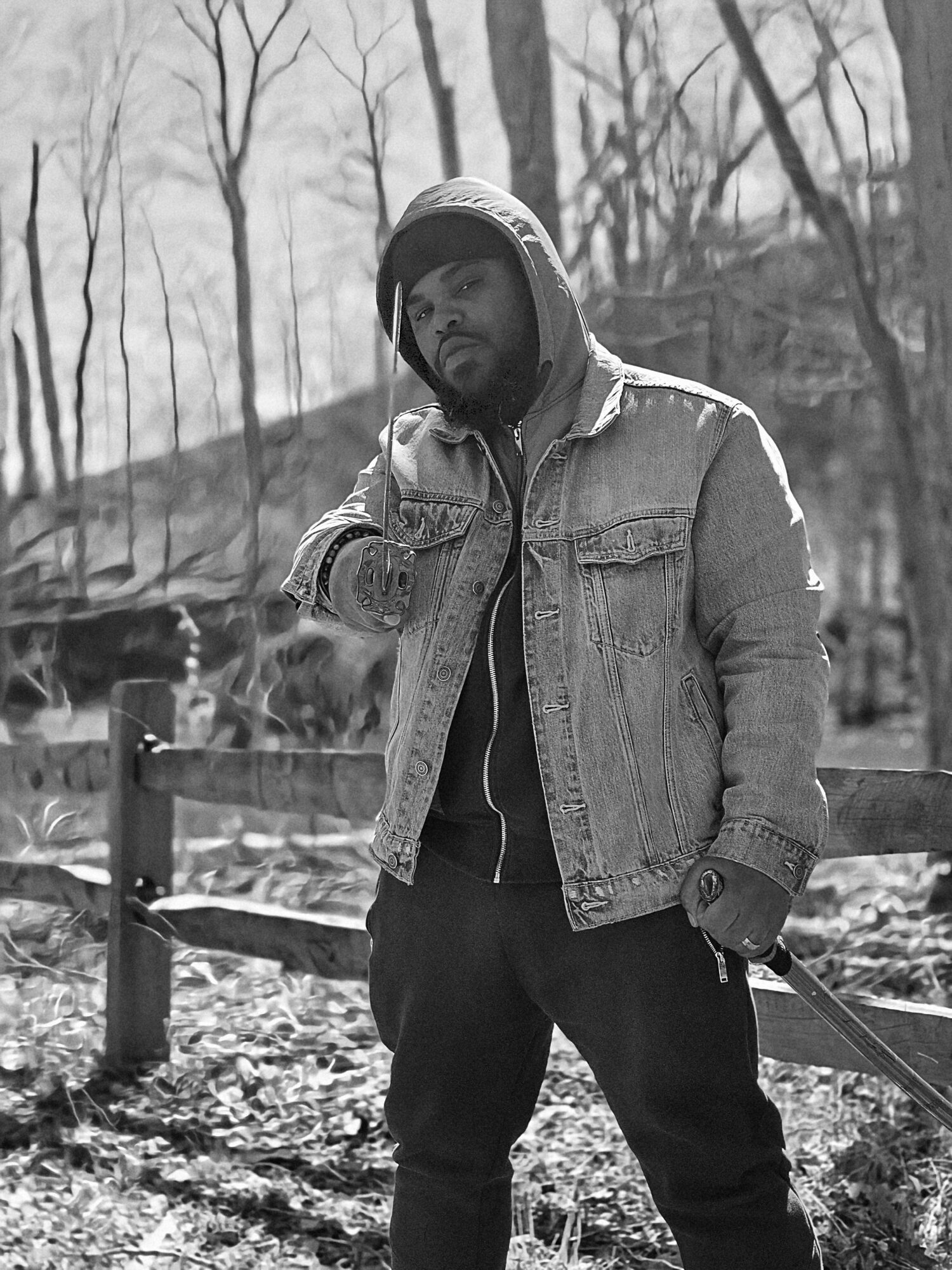 Man in denim jacket and hoodie standing outdoors near a wooden fence, trees in background, black and white photo.