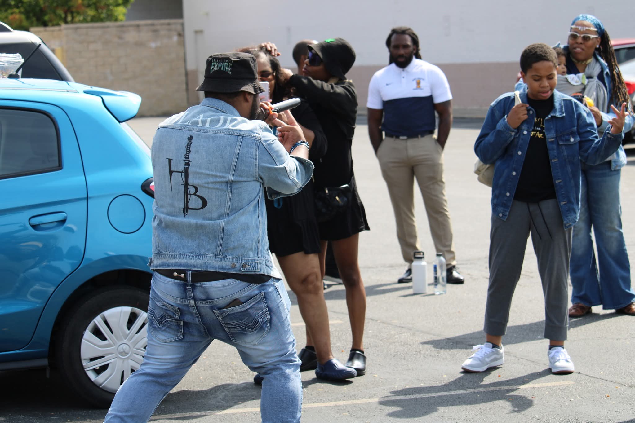 Group of people standing outdoors near a blue car, some talking and others looking around, with a white wall in the background.