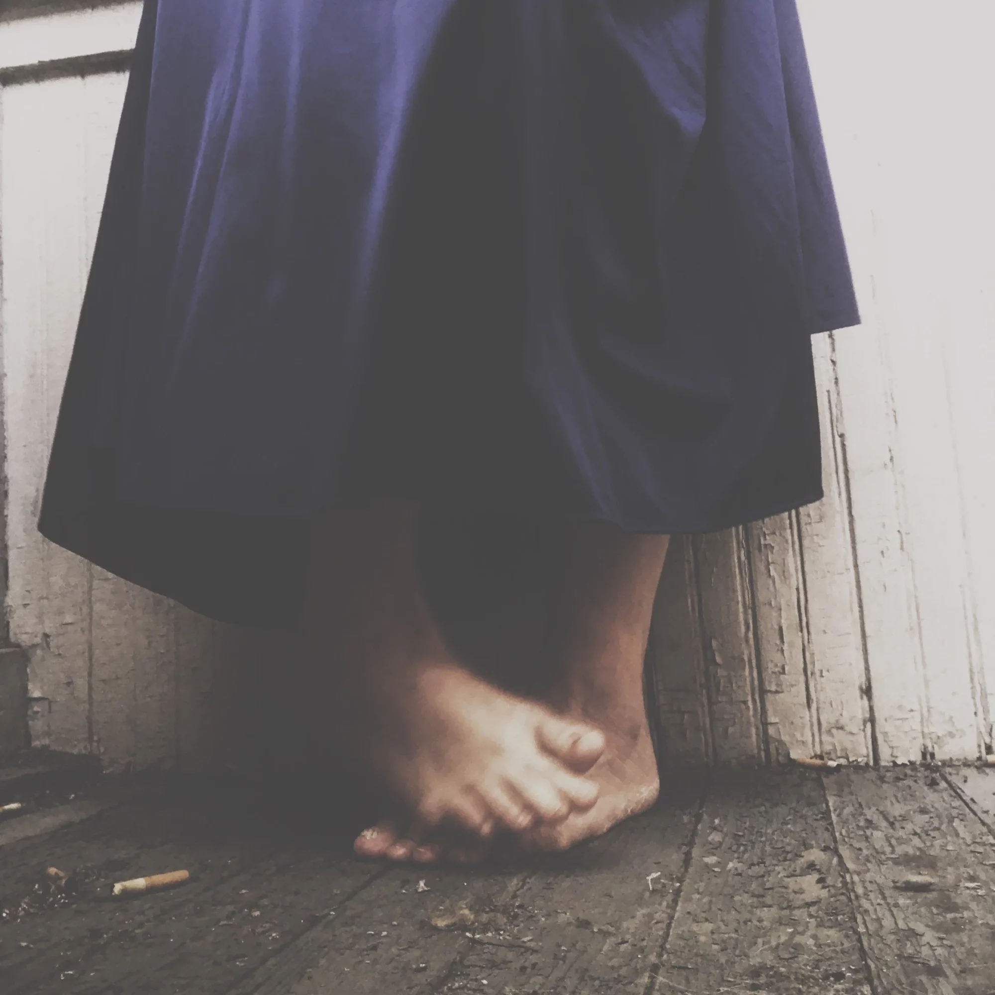 Person standing barefoot on a wooden floor, wearing a dark dress, against a white wooden wall.