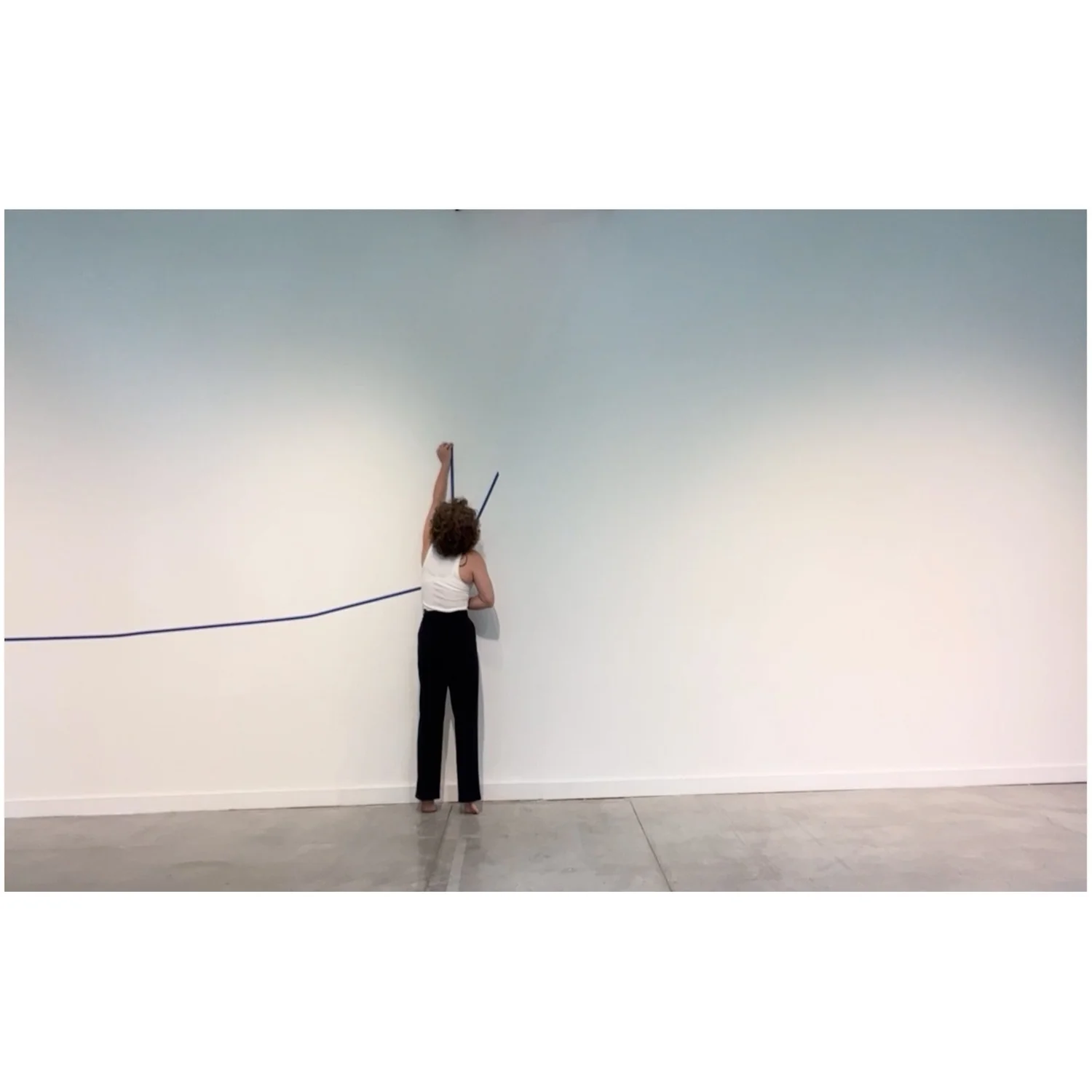 Person standing with back to camera, reaching up to a blank wall, holding a paintbrush or similar object, with a cord trailing behind.