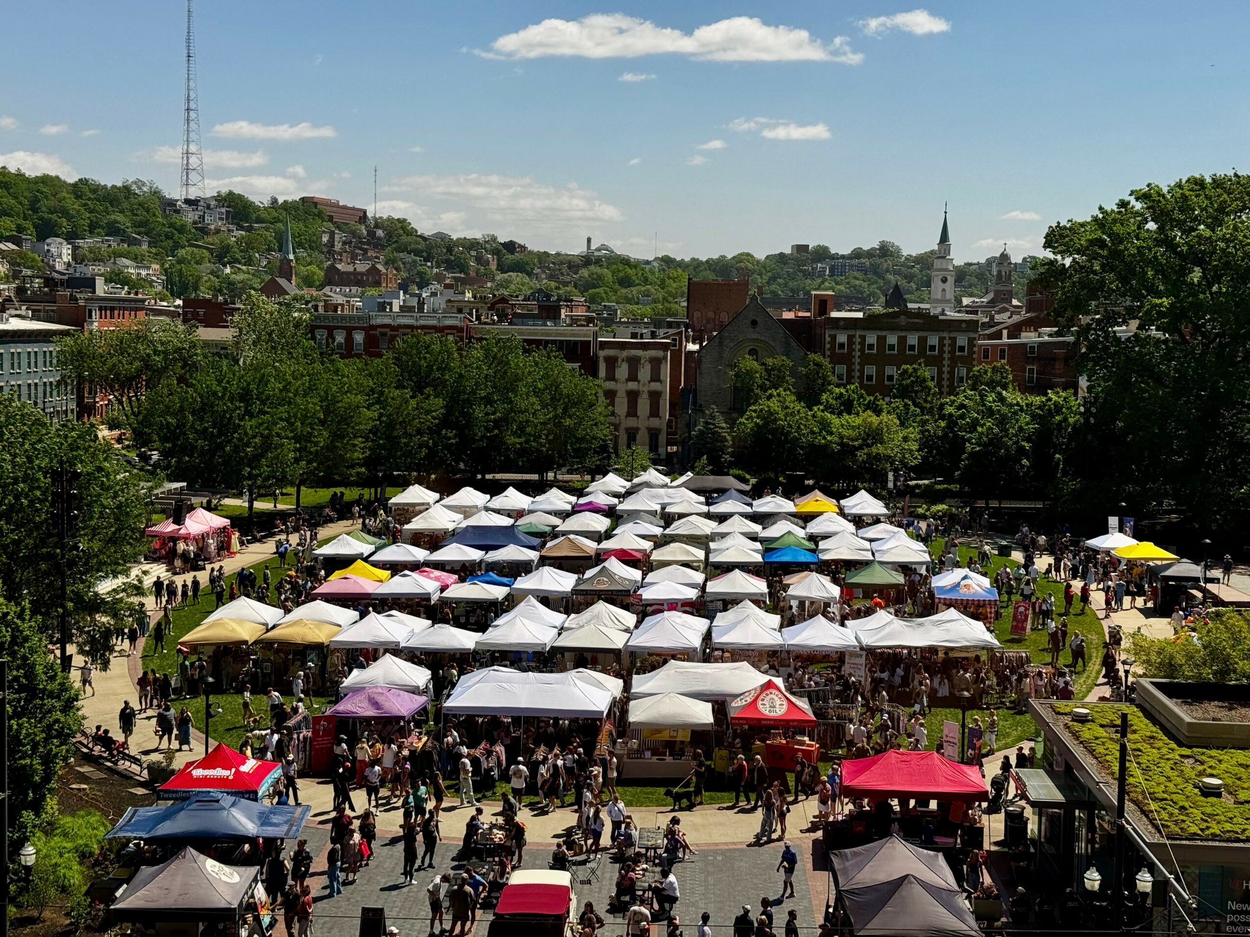 Outdoor market with numerous tents and people, surrounded by trees and city buildings, under a partly cloudy sky.