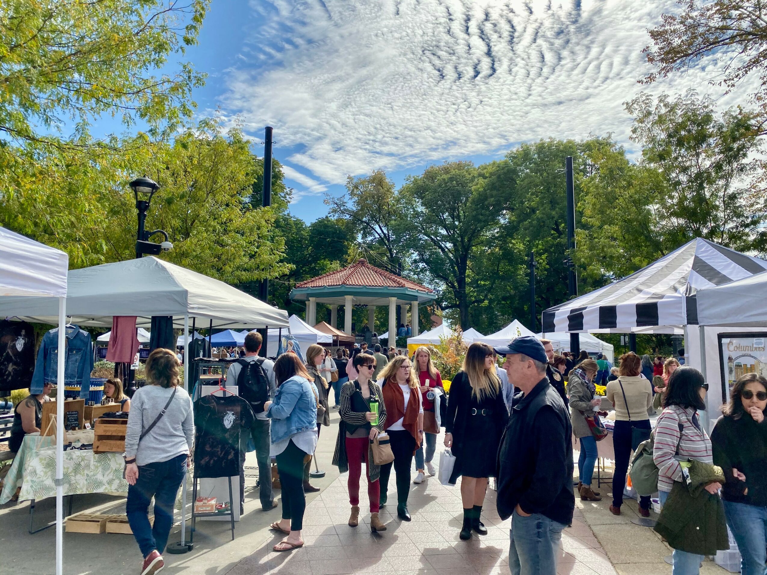 People browsing outdoor market stalls under tents, trees, and a gazebo with a partly cloudy sky.