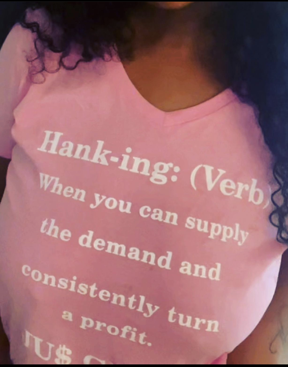 Person wearing a pink shirt with white text about supply and demand, curly hair visible on the side.
