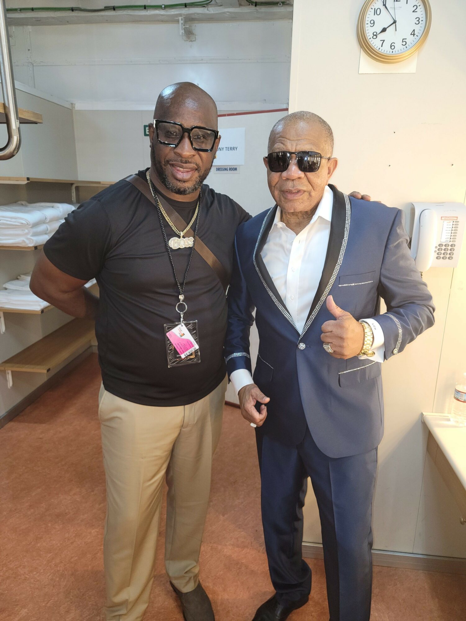 Two men standing indoors, one in a suit and sunglasses, the other in a black shirt and khakis, both smiling.