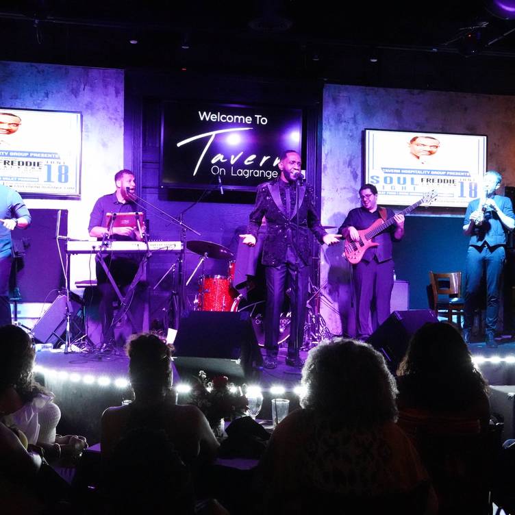 Musicians perform on stage with a singer, keyboard, guitar, and microphone, in a dimly lit venue with audience silhouettes.