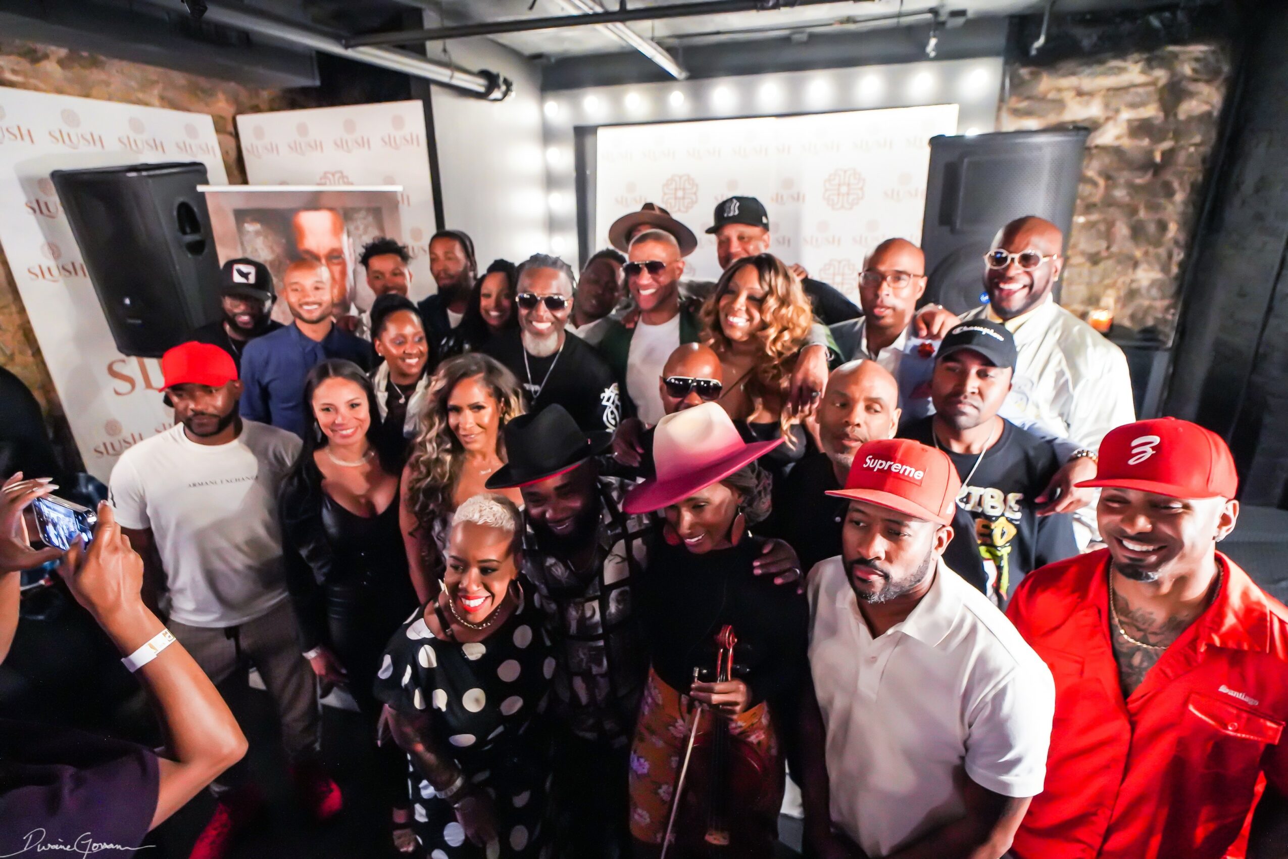 Group of people gathered indoors, some wearing hats and sunglasses, smiling and posing for a photo.