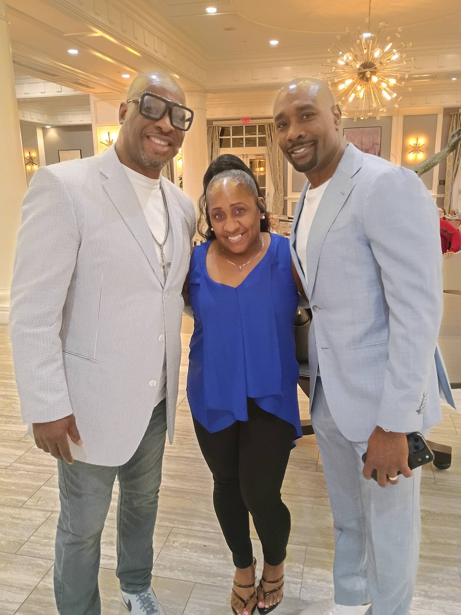 Three people smiling indoors, two men in light suits and a woman in a blue top, standing close together.