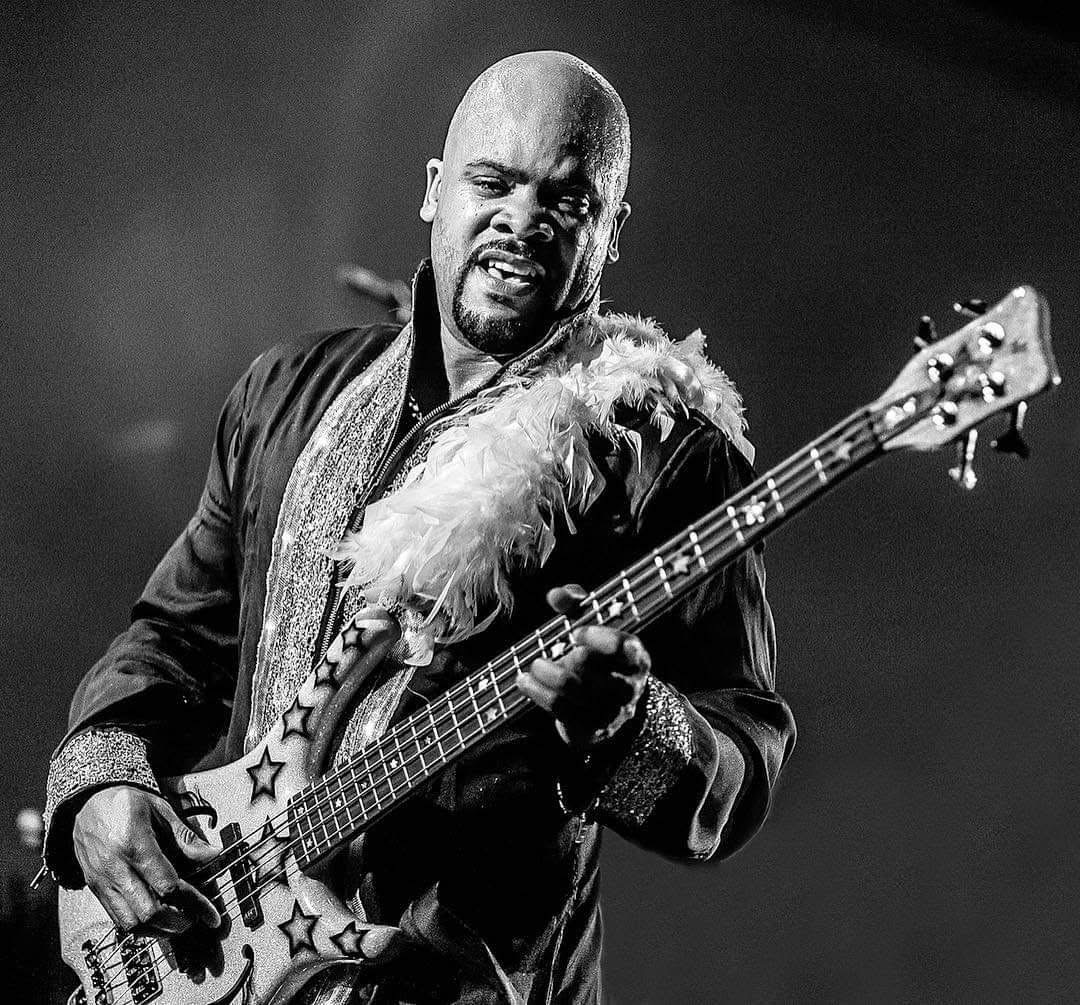 Bald man playing a guitar with a feathered strap, wearing a dark jacket, on stage in black and white.