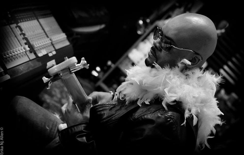 Person with bald head, glasses, and feathered clothing, holding a microphone in a dark setting.