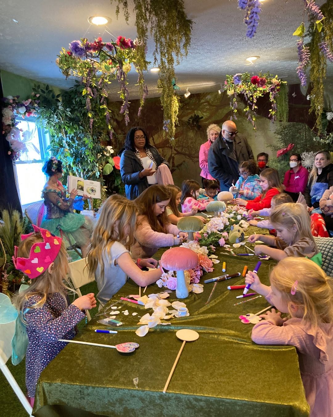 Children and adults gather around a table with craft supplies, decorated with hanging flowers and greenery, in a festive indoor setting.