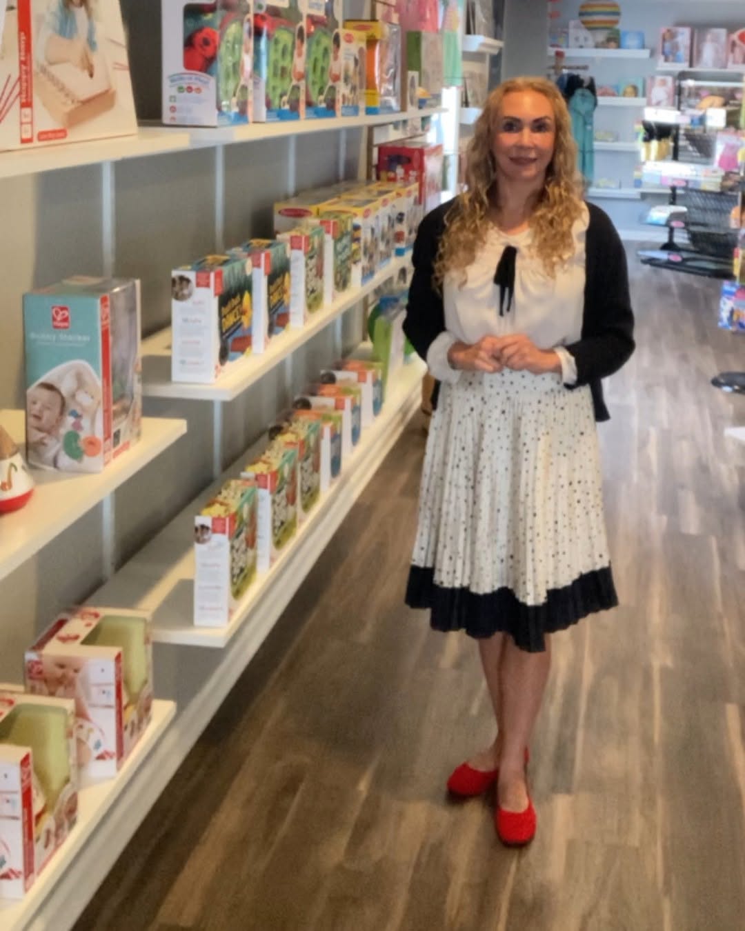Woman with curly blonde hair in a white dress, black cardigan, and red shoes standing in a bookstore aisle with books on shelves.