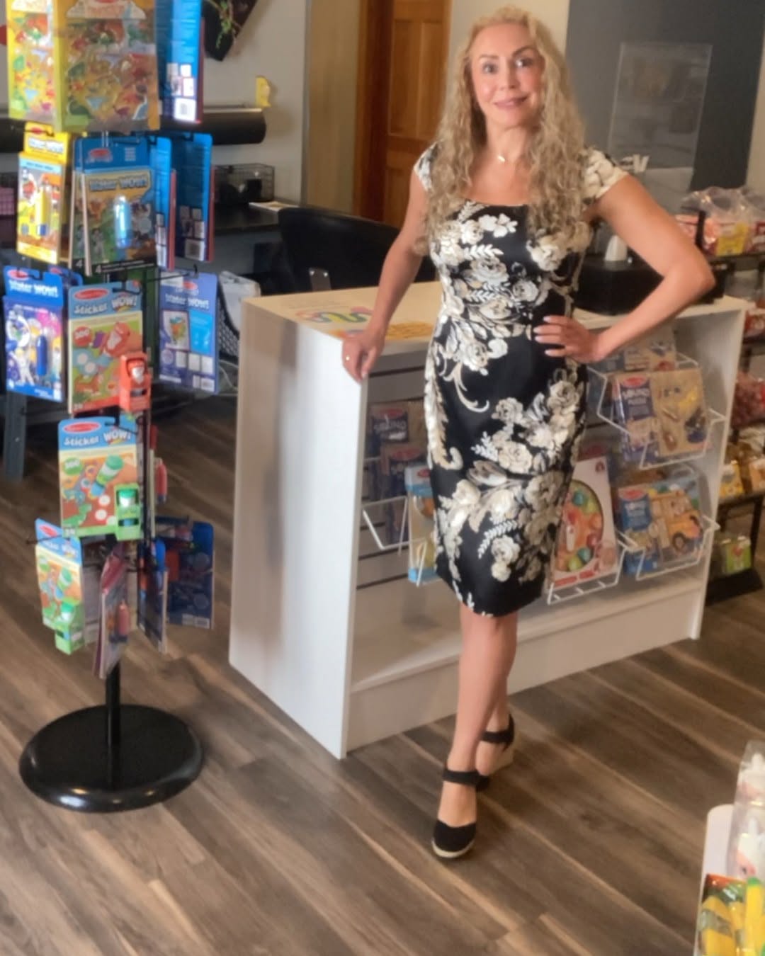 Woman with long curly blonde hair in a black and white floral dress, standing in a toy store, smiling, with toys on shelves behind her.