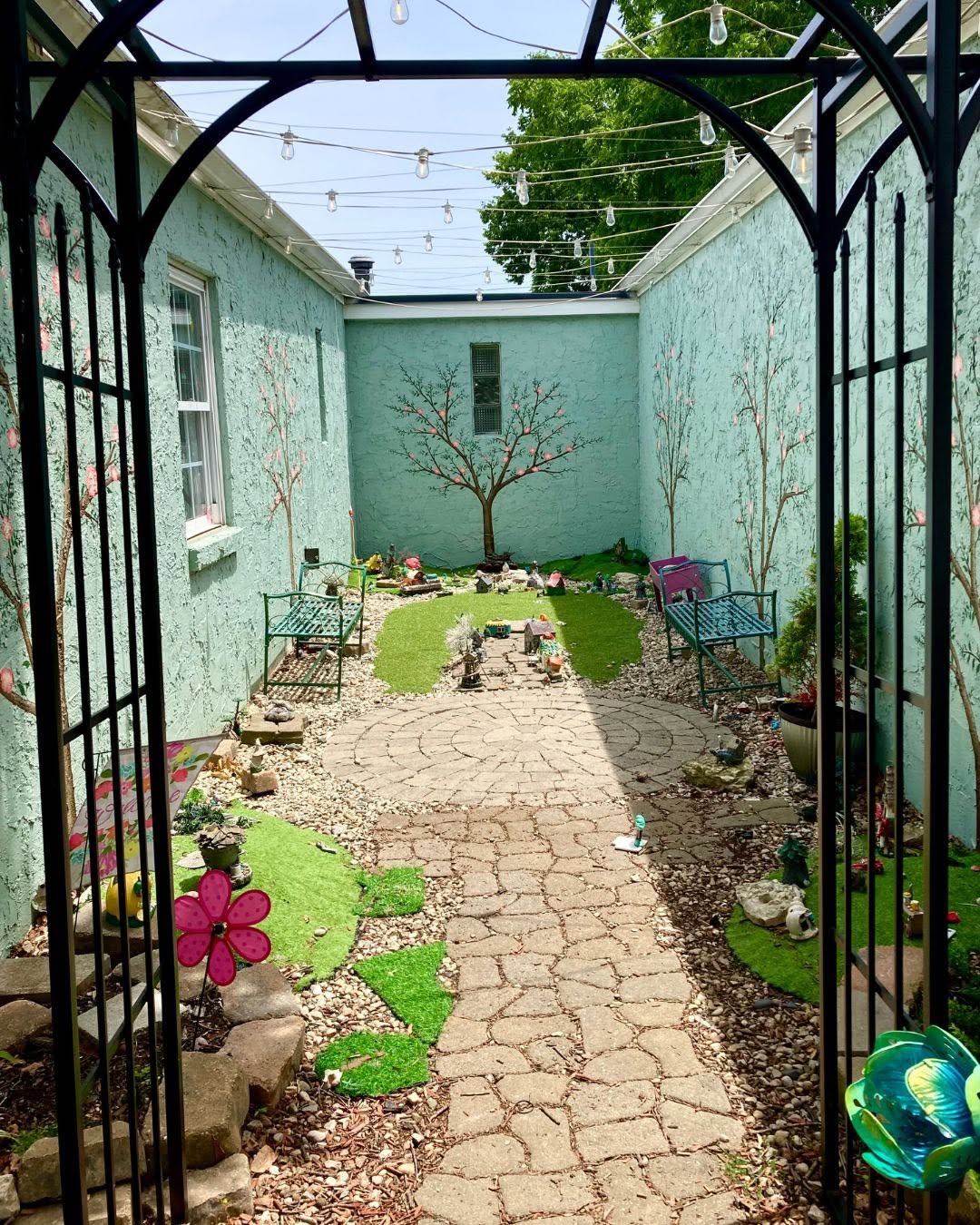 View through a black metal gate into a small garden with a stone pathway, potted plants, and a painted wall with trees.