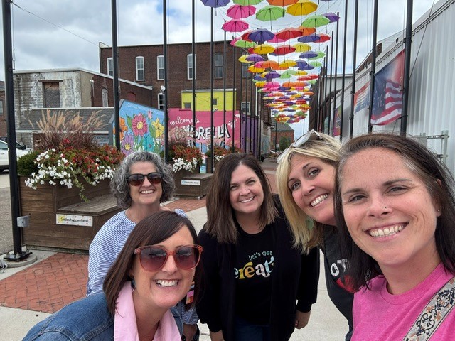 Five women smiling outdoors under colorful umbrellas hanging overhead, with buildings and flower planters in the background.