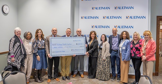 Group of people standing indoors, holding a large check, in front of a wall with 'AULTMAN' logo, smiling.