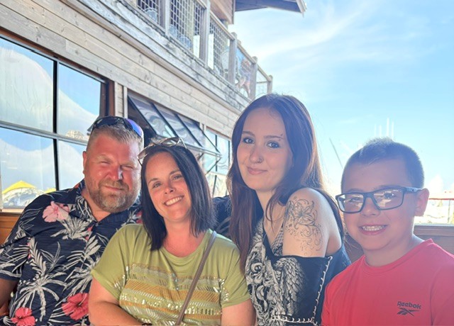 Four people smiling outdoors near a building with large windows, sunny sky in background.