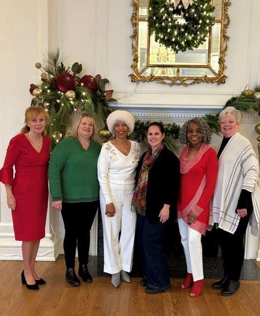 Six women standing in front of a decorated fireplace with a mirror and Christmas decorations, smiling together.