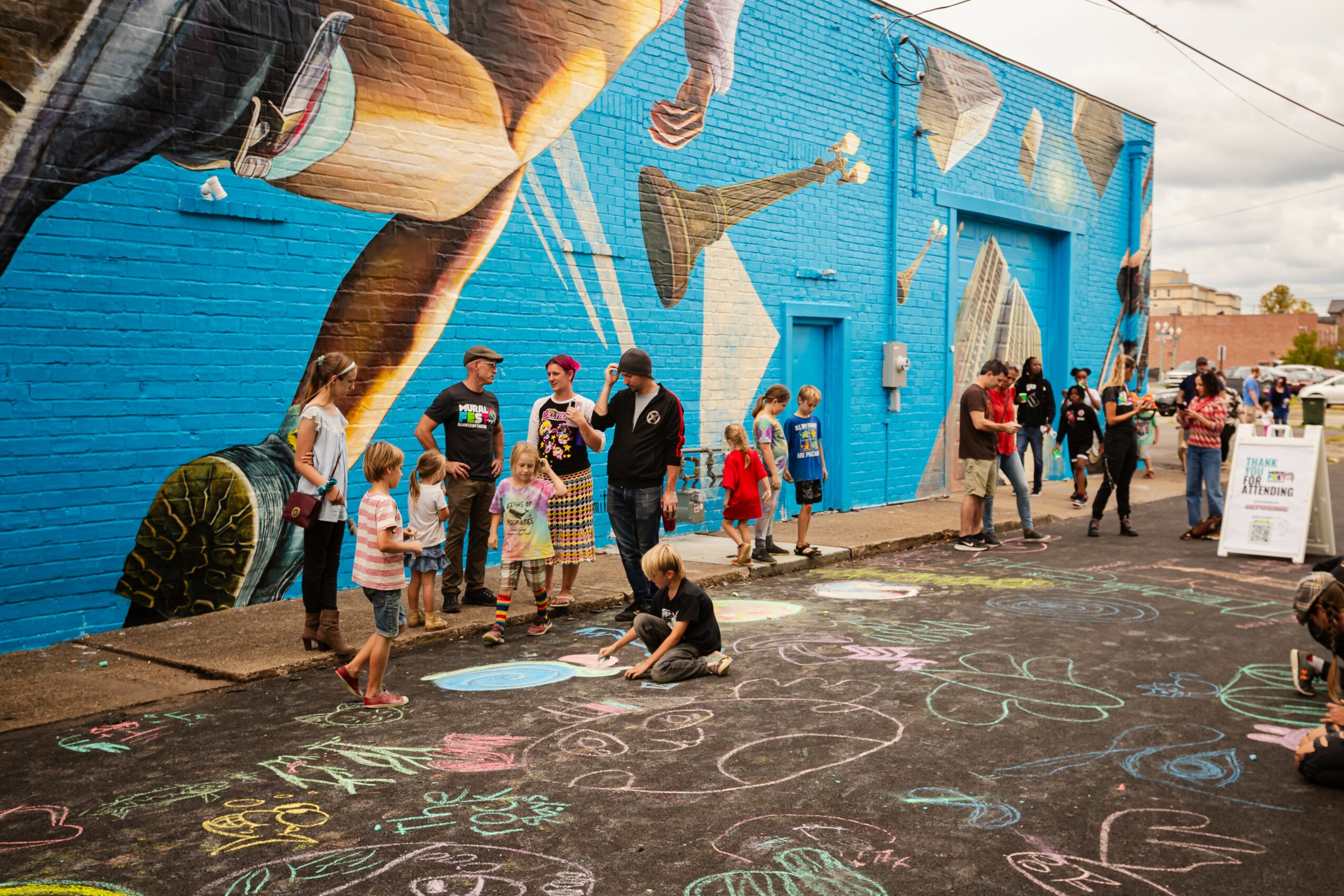 People, including children, draw with chalk on a sidewalk in front of a blue mural wall with artwork and guitars.