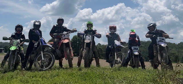 Group of seven people with motorcycles standing on grass under cloudy sky, some wearing helmets, in outdoor setting.