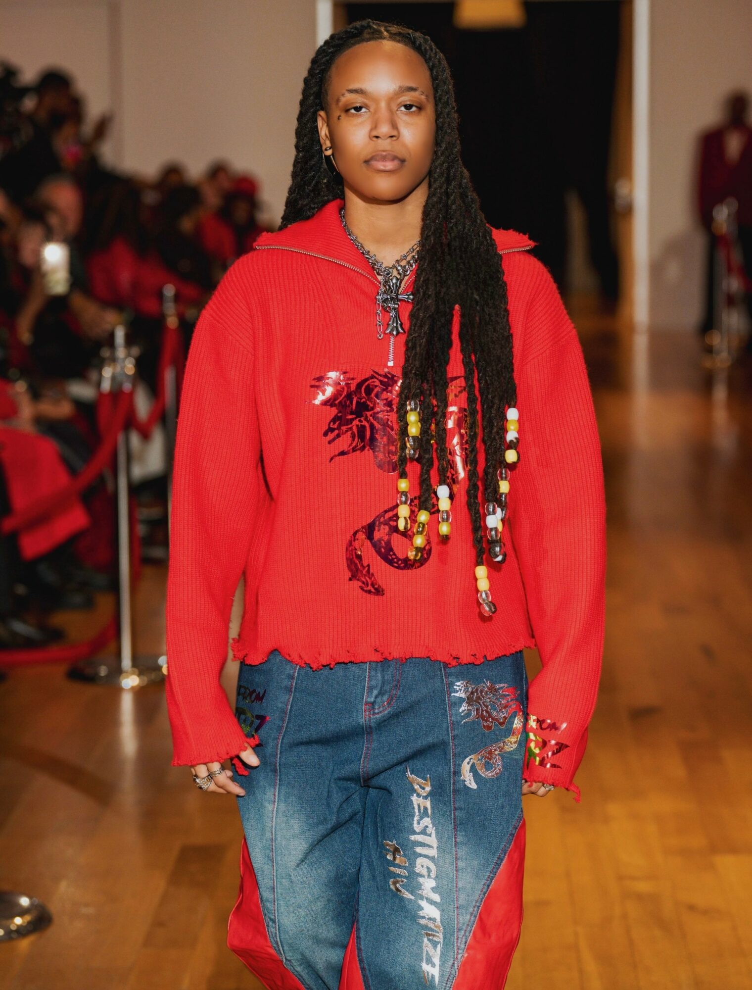 Young woman walking on runway in red hoodie with dragon design, blue jeans, braided hair, audience in background.