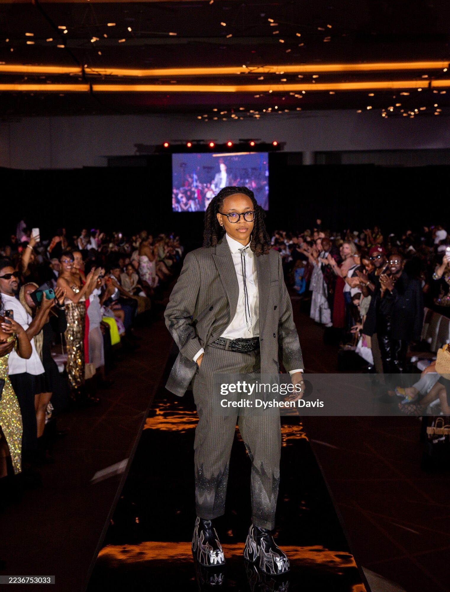 Person in a suit walking on a runway at an event with a large audience and a screen in the background.