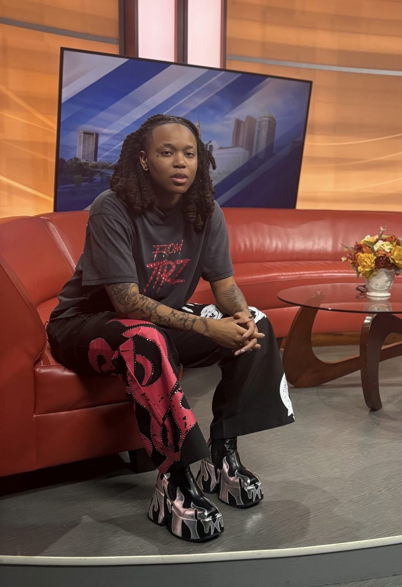 Person with long hair sitting on a red couch in a TV studio, with a large screen behind them and a flower arrangement on a table.