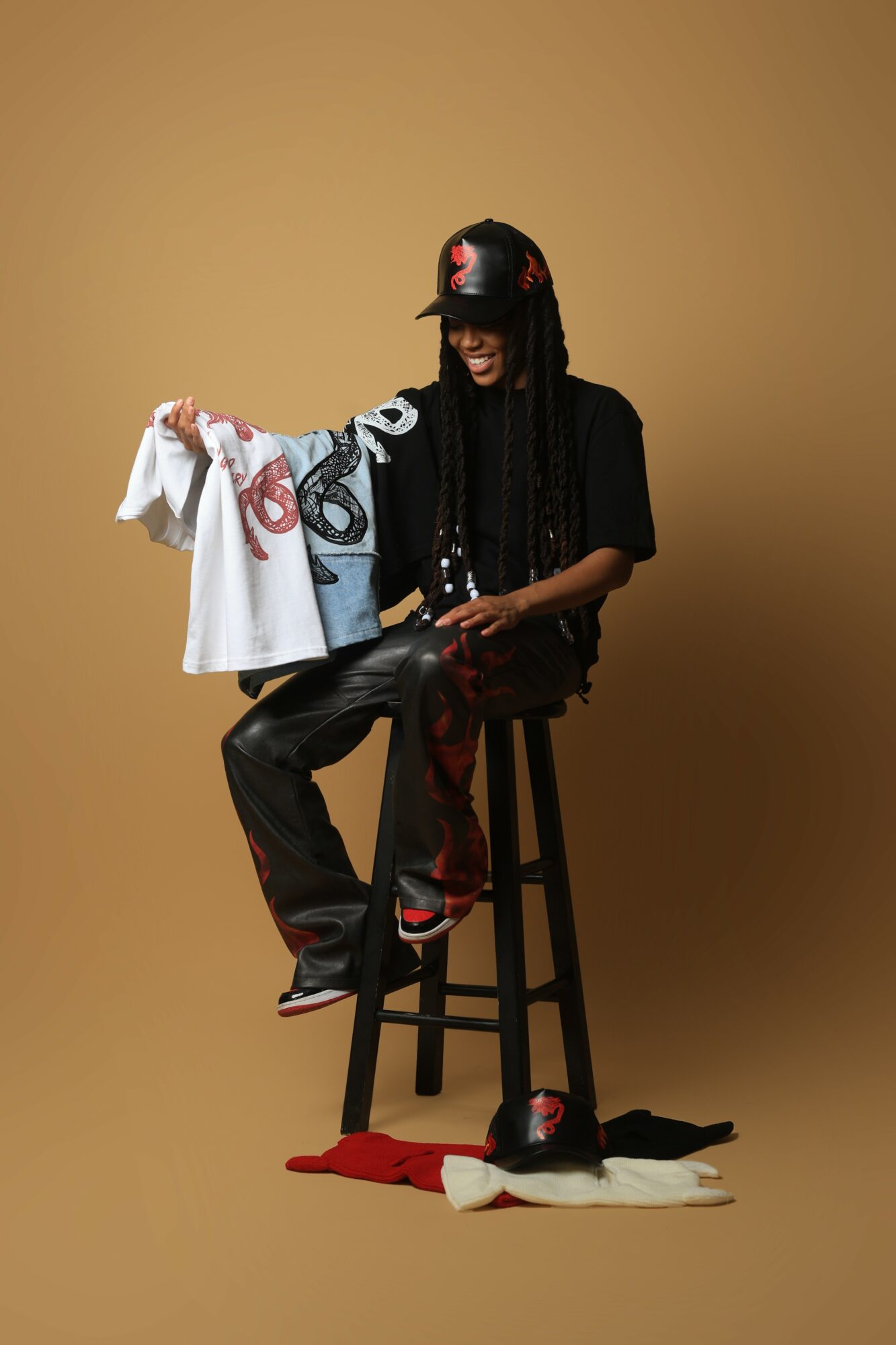 Person sitting on a stool holding a towel, wearing a mask, with clothing and items on the floor, against a plain background.