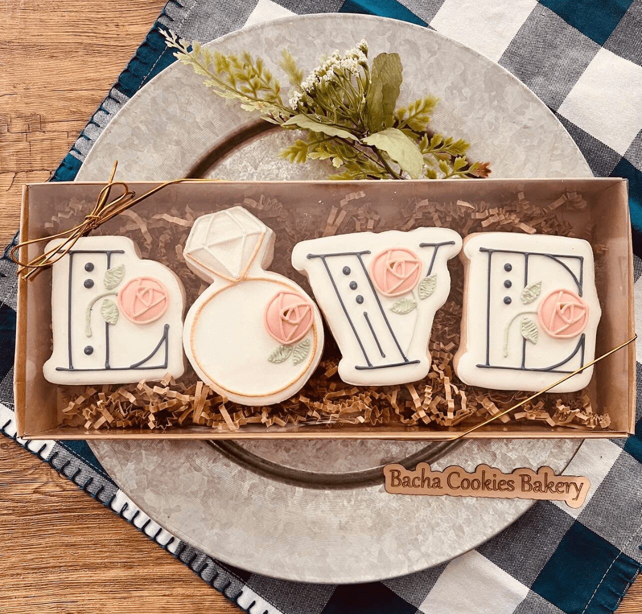 Decorative cookies spelling 'LOVE' with pink roses, arranged in a box on a plate, with a checkered cloth background.