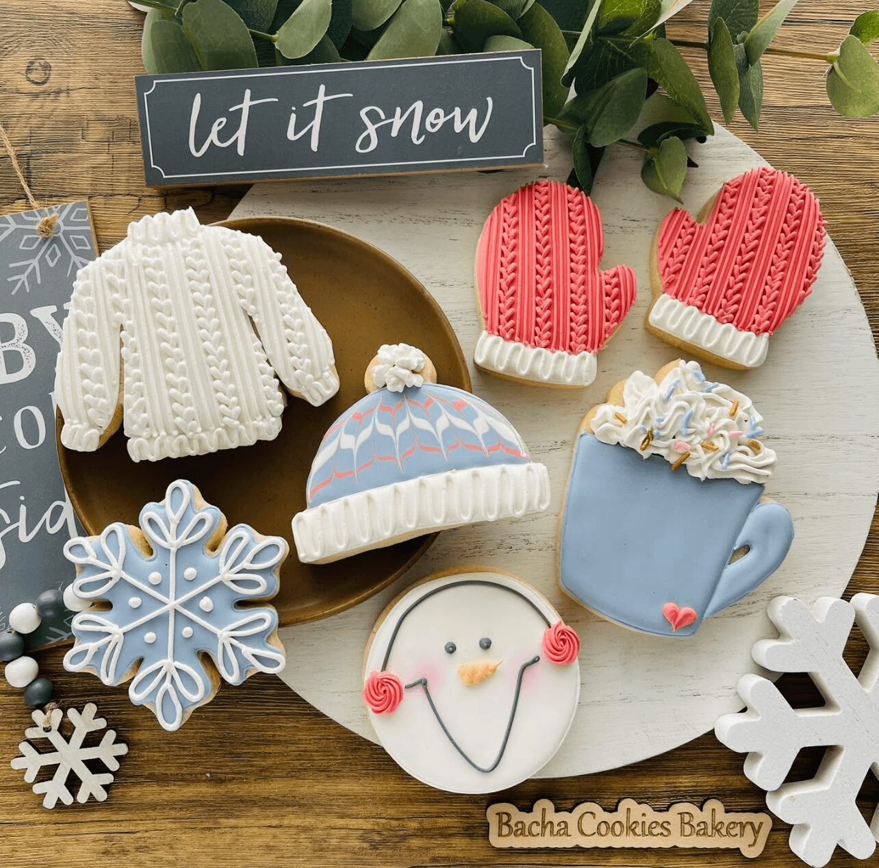 Assorted winter-themed cookies including a sweater, mittens, snowflake, hat, snowman, and mug, on a white tray with a sign and greenery.