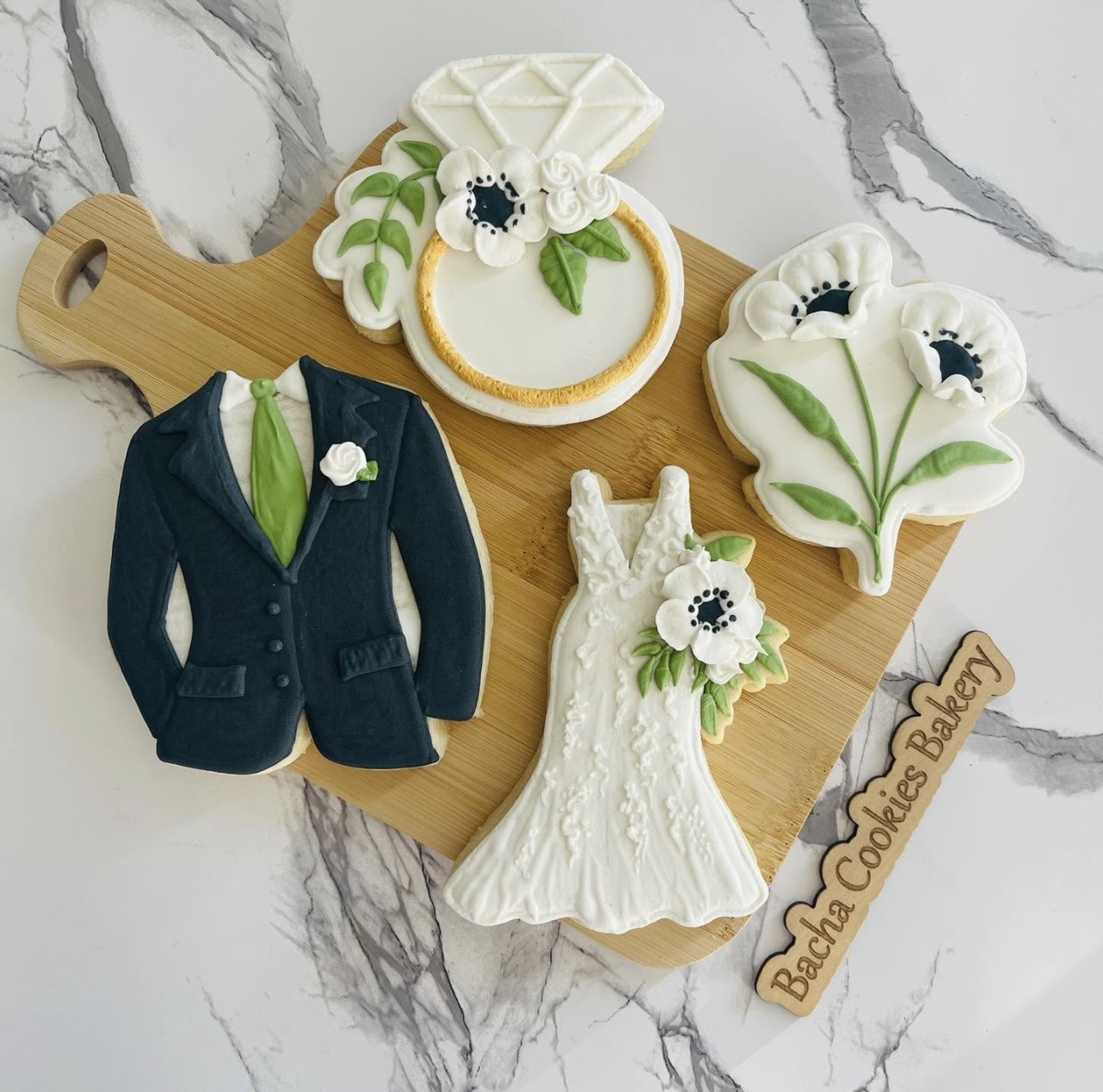 Decorative wedding-themed cookies and a suit cookie on a wooden board, with a marble surface background.