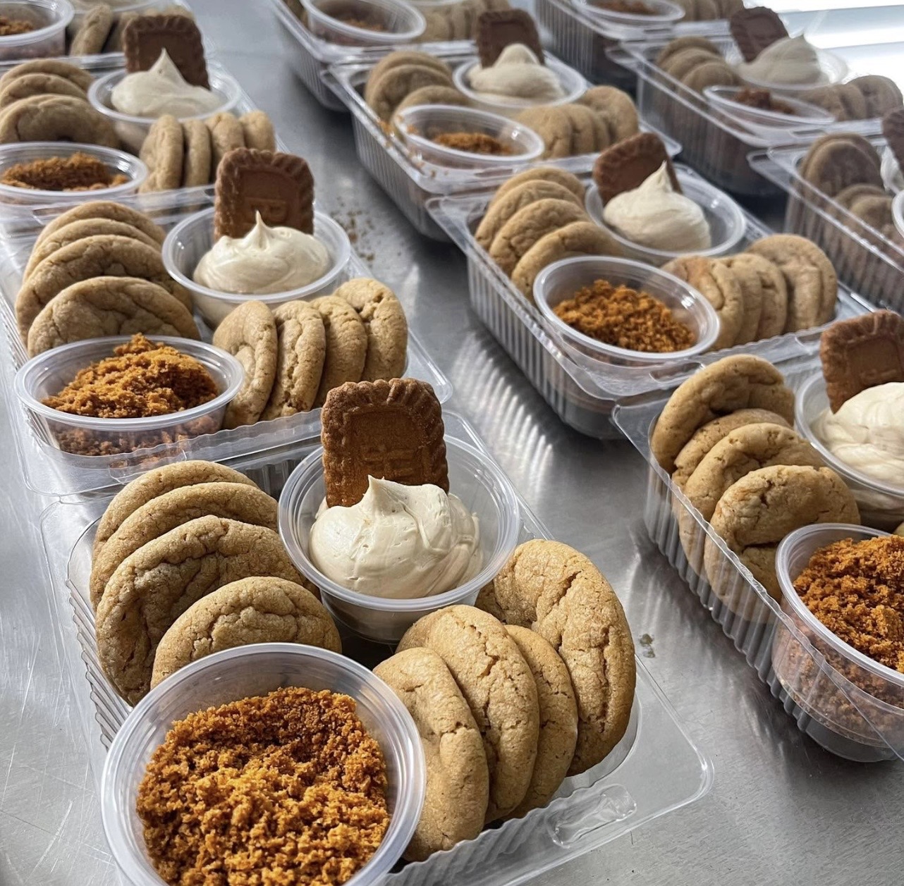 Multiple clear plastic containers filled with cookies, whipped cream, and caramel topping, arranged on a metallic surface.