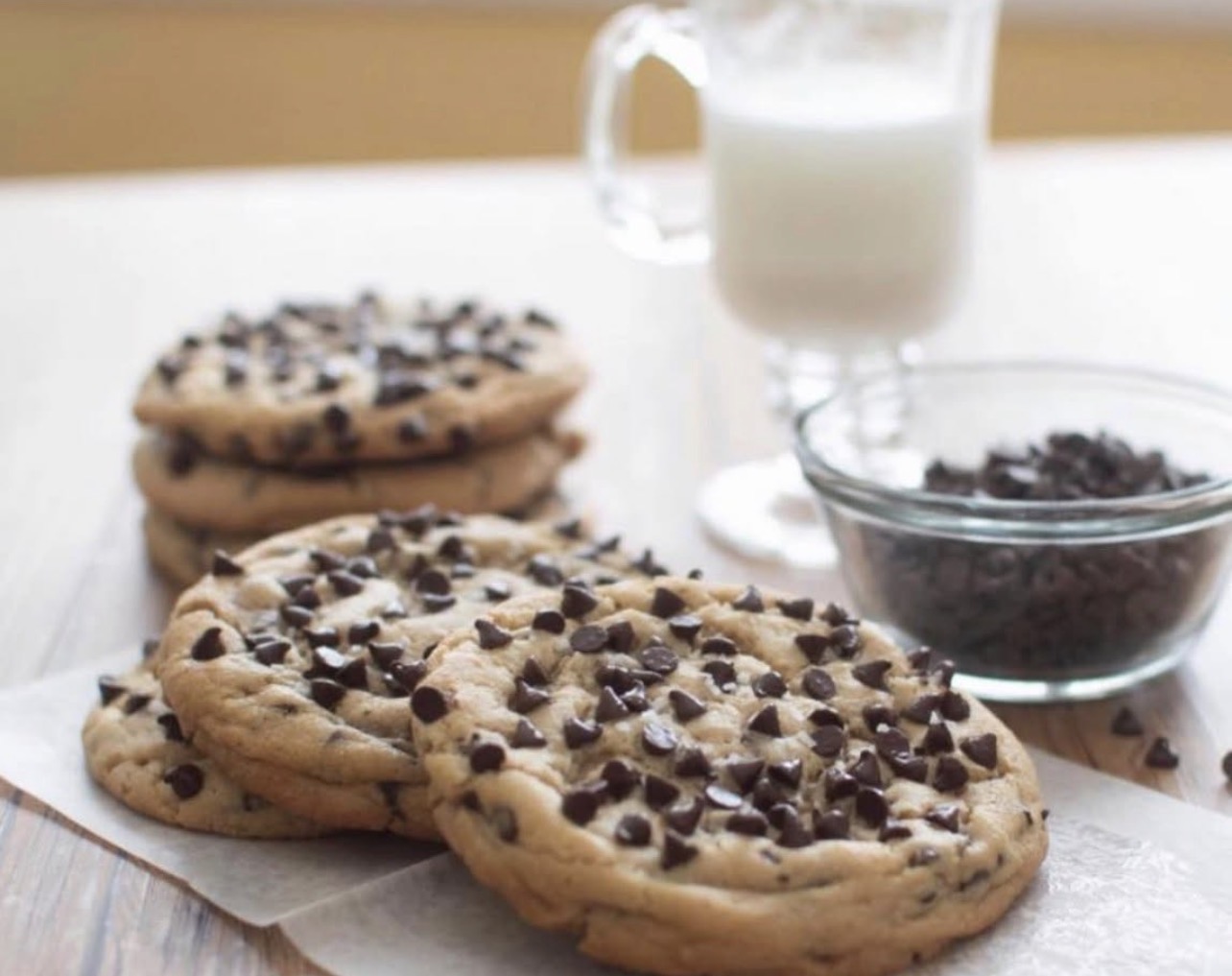 Three chocolate chip cookies on paper, a glass of milk, and a bowl of chocolate chips on a table.
