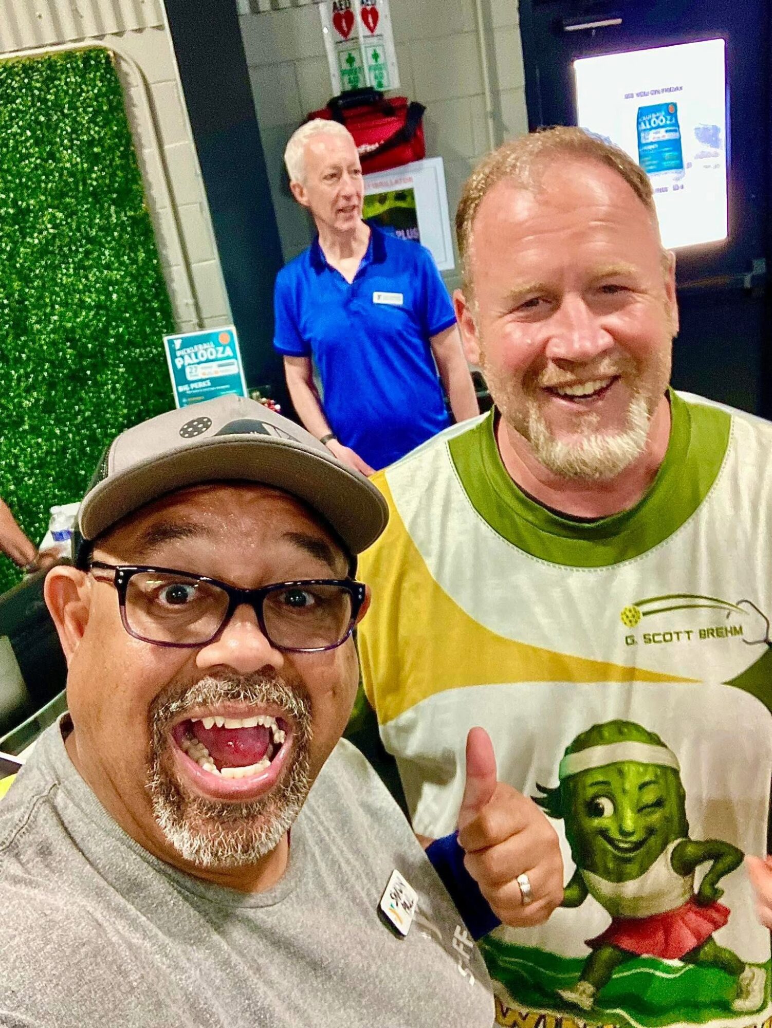 Two smiling men taking a selfie, one giving a thumbs-up, with a woman in a blue shirt in the background.