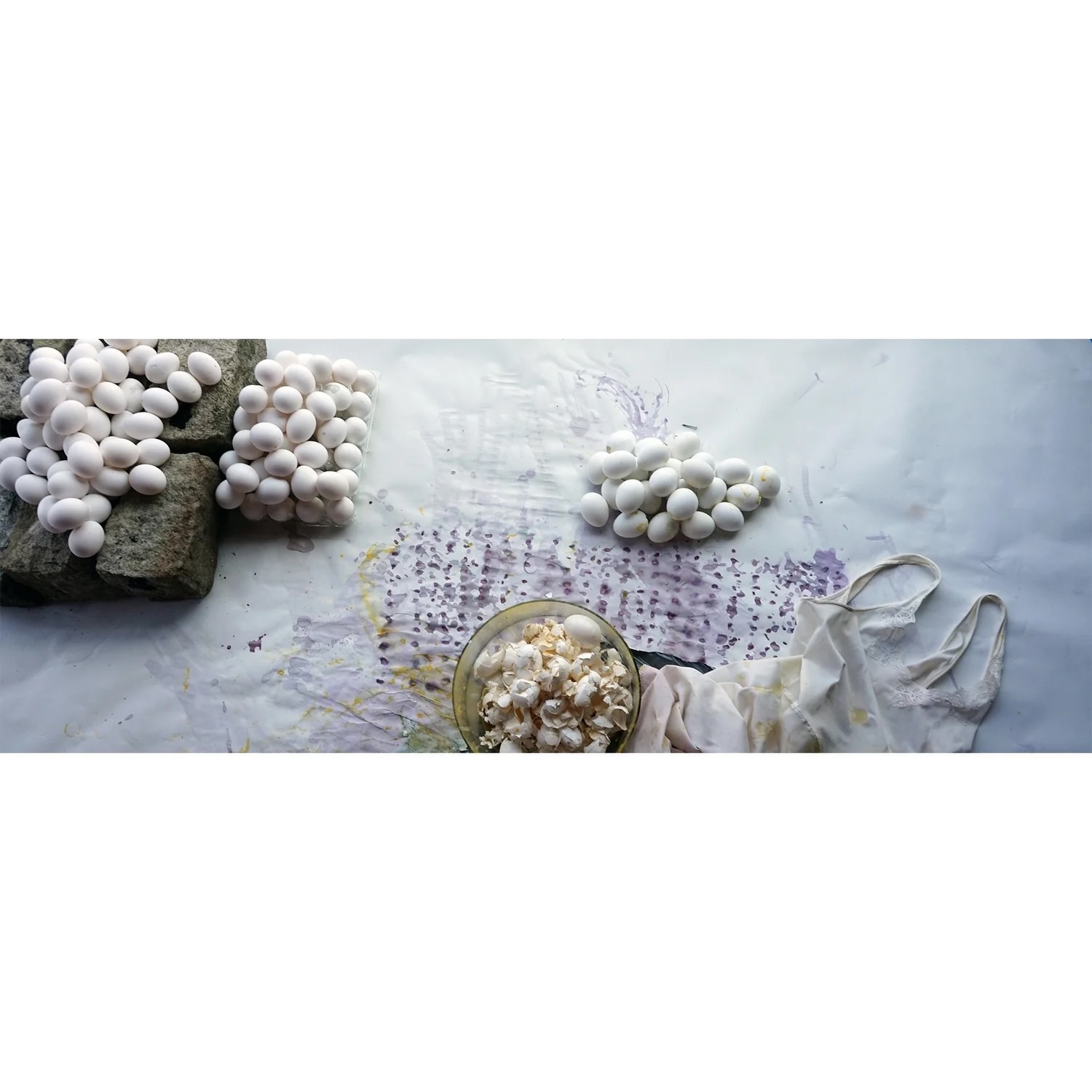 White eggs and stones on a white cloth with a bowl of eggs and a ribbon, viewed horizontally.