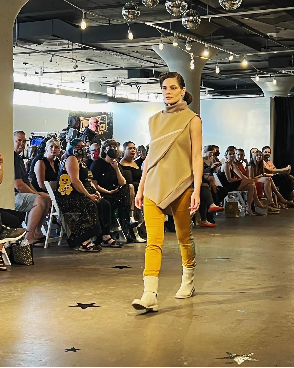 Model walking on runway in beige top and yellow pants, audience seated in background, indoor fashion show setting.