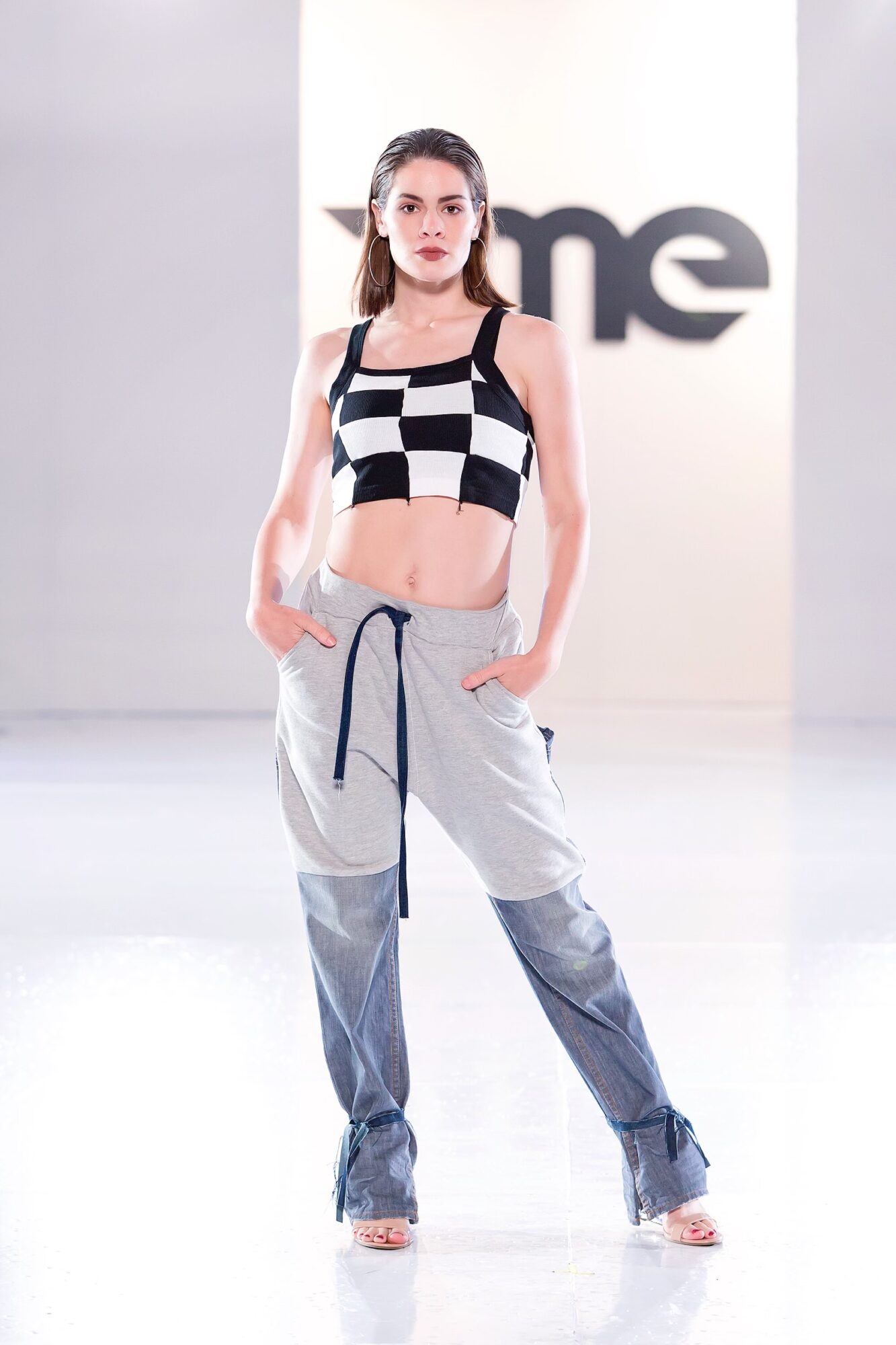 Young woman walking on runway, wearing checkered crop top and gray pants with hands in pockets, in a bright studio.