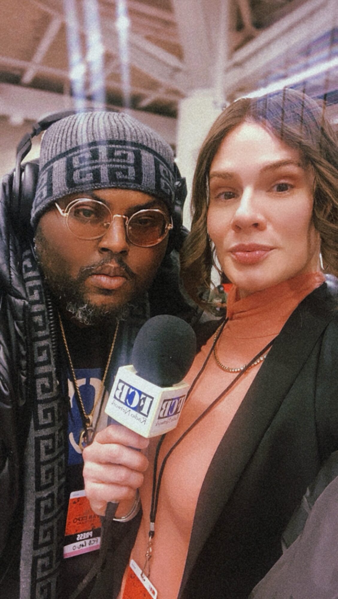 Two people, one holding a microphone, pose for a photo indoors with a high ceiling and lighting fixtures.