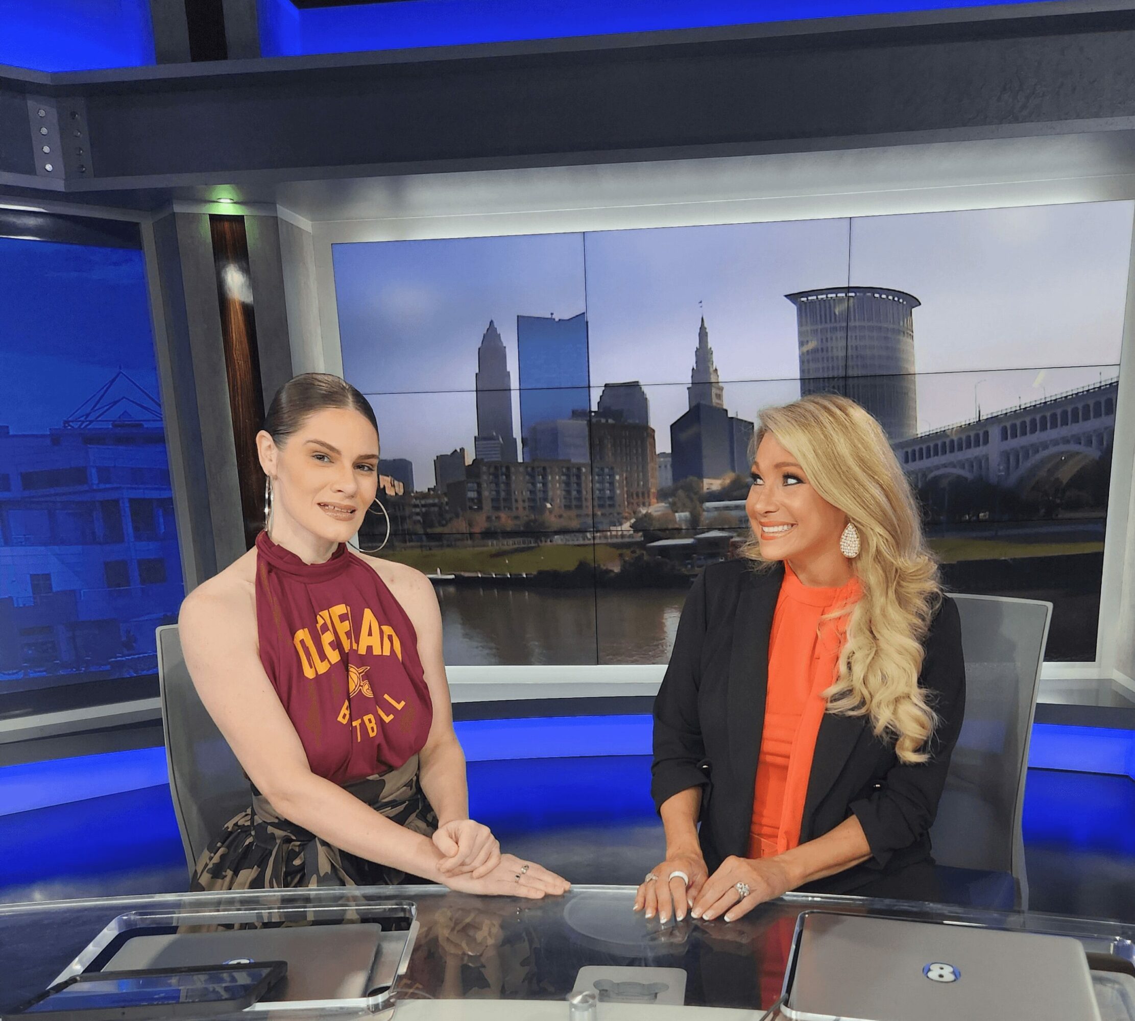 Two women sit at a news desk with a city skyline background on a large screen behind them.