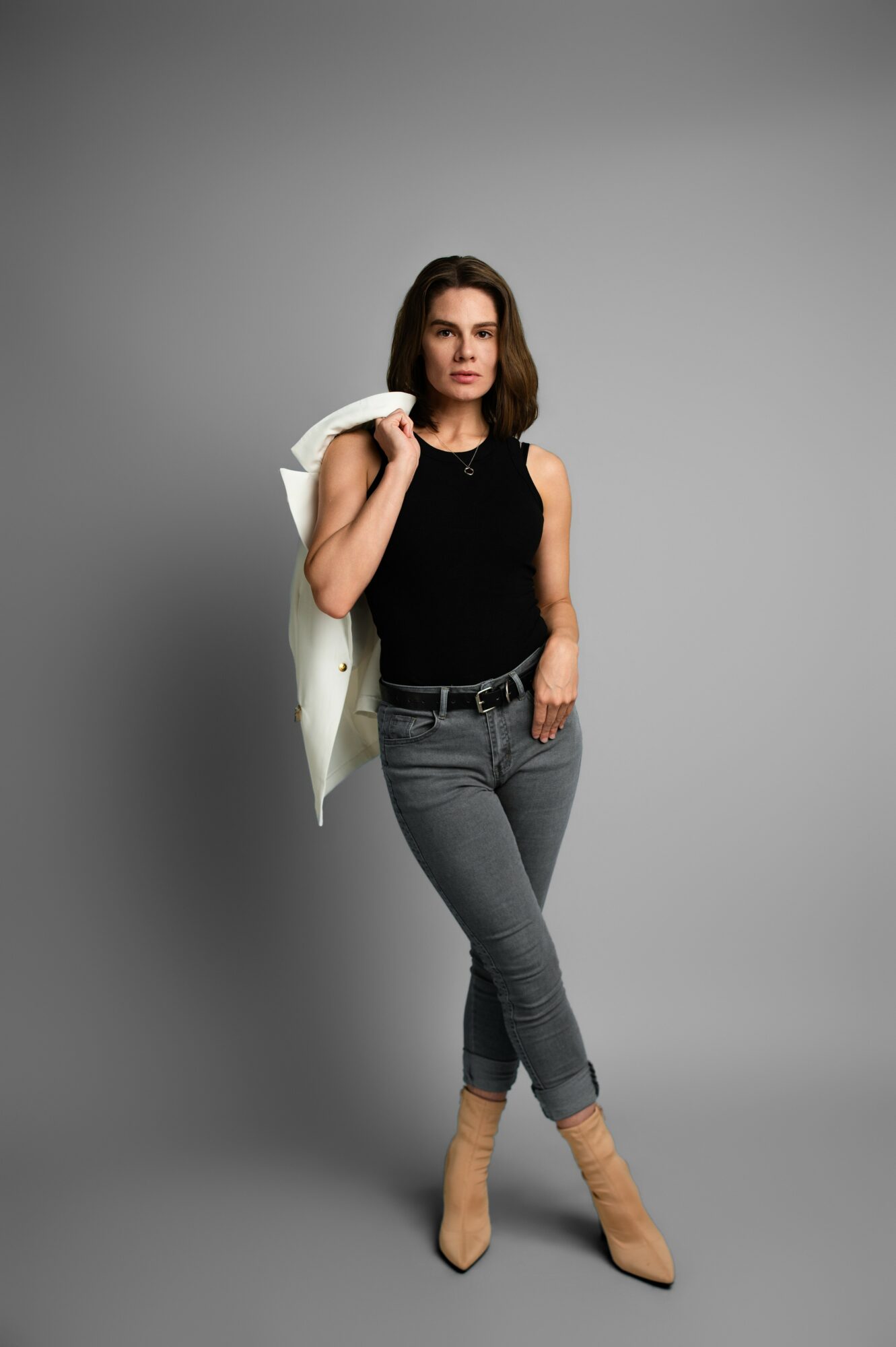 Woman with shoulder-length hair in a black sleeveless top and gray jeans, holding a white jacket over her shoulder, standing against a gray background.