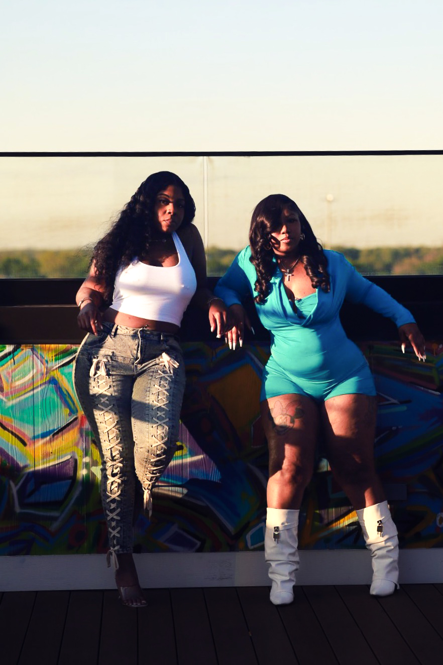 Two women with long dark hair standing outdoors near a colorful graffiti wall, one in a white top and patterned pants, the other in a blue outfit.