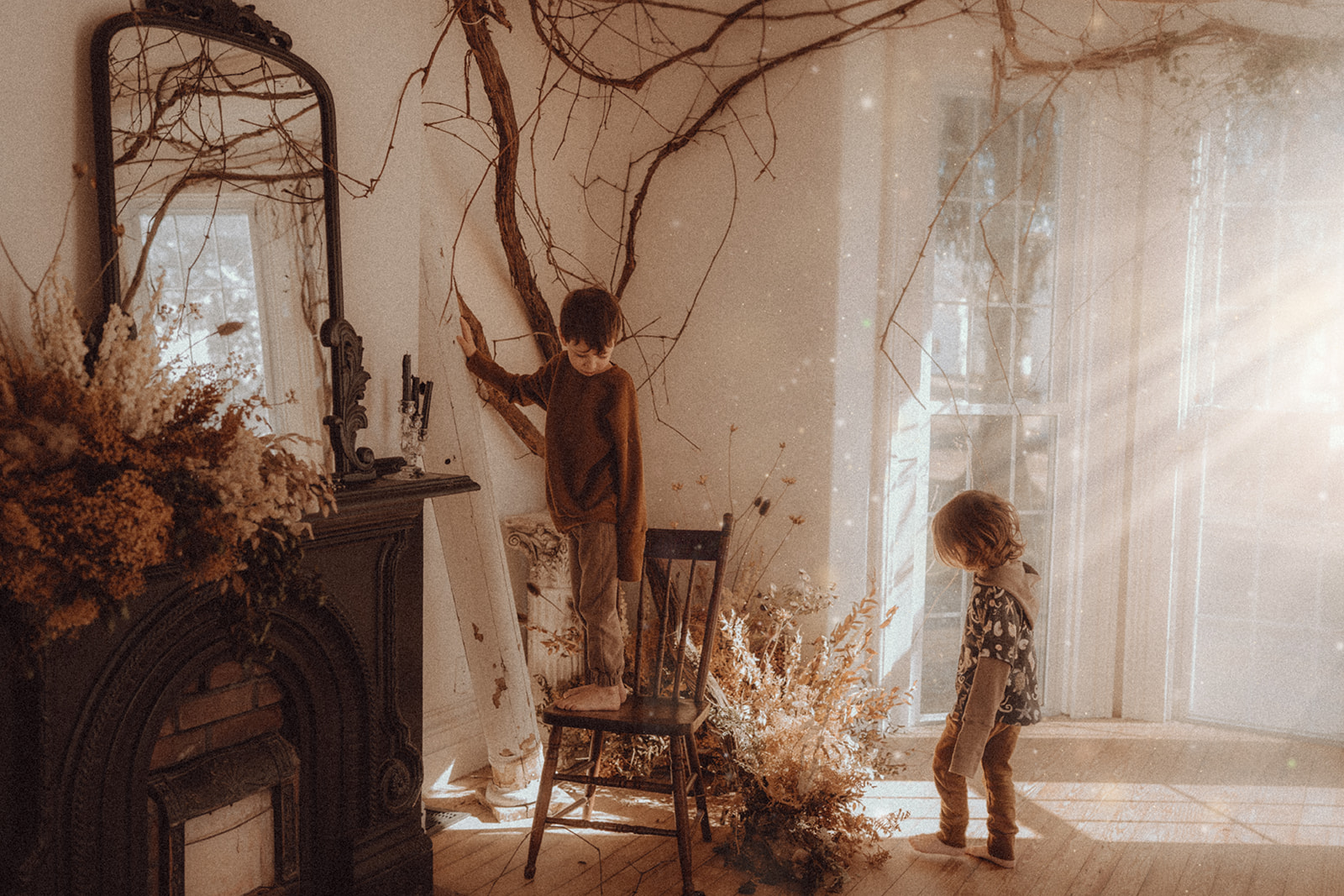 Two children in a room with sunlight streaming through large windows, one reaching for a branch on the wall, the other standing nearby.