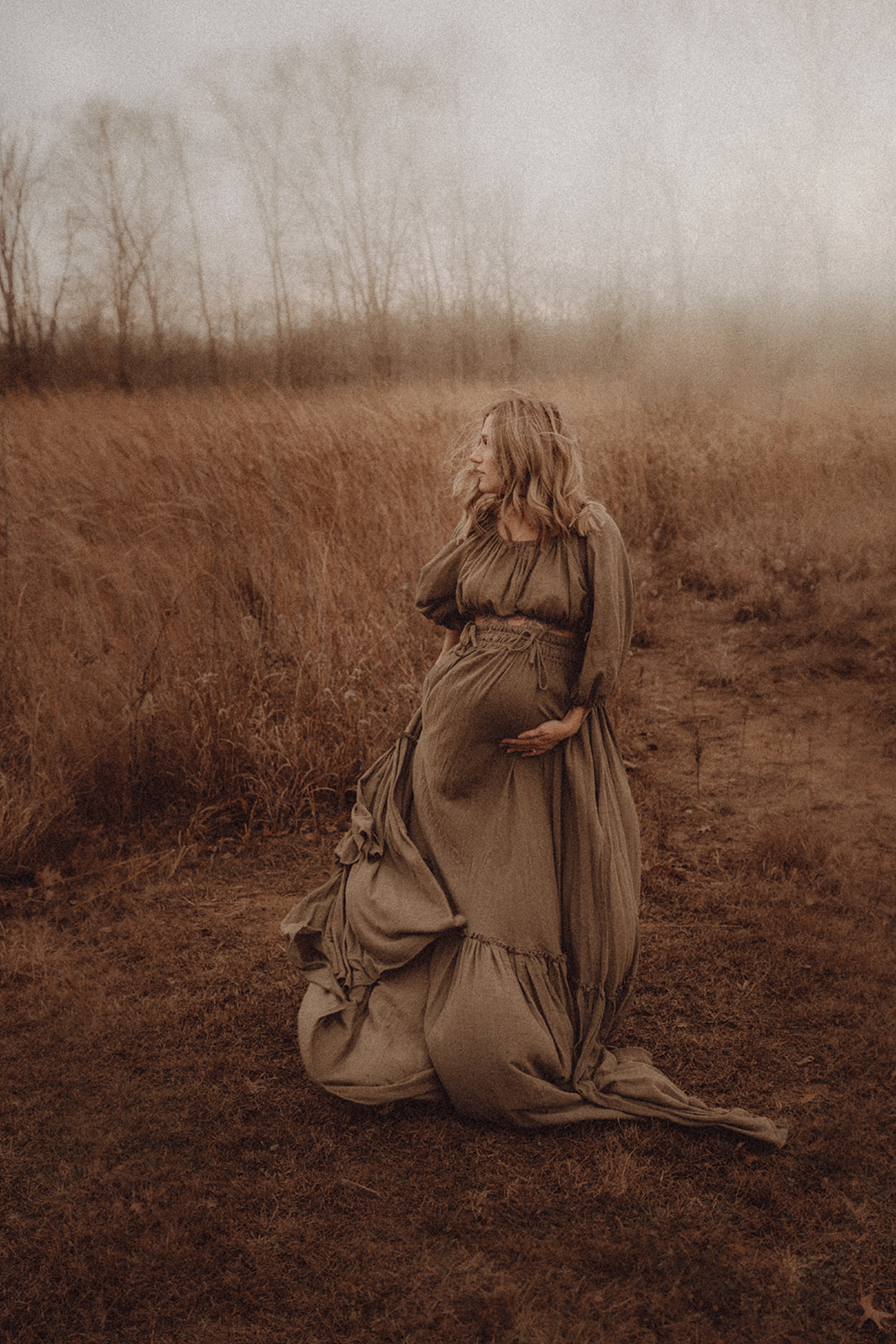Woman in a long dress standing in a field with tall grass and trees in the background.