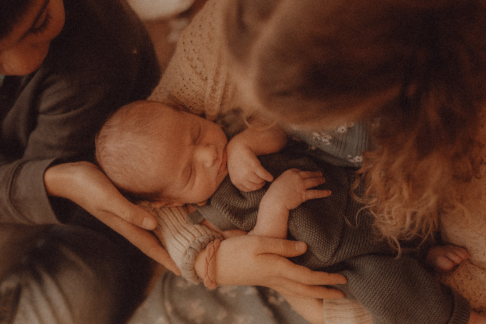 Baby lying in adult's arms, surrounded by people, with a woman with curly hair nearby, in a close setting.