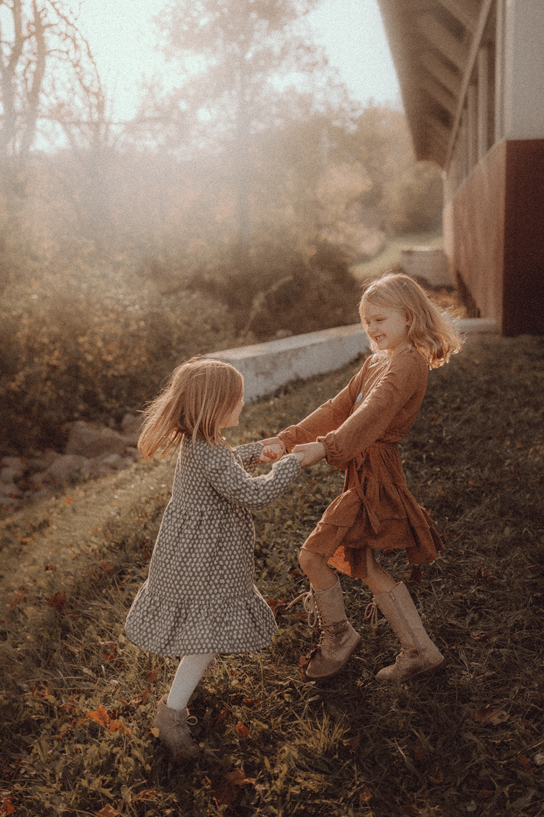 Two young girls holding hands and playing outdoors on a grassy slope near a house, with trees in the background.