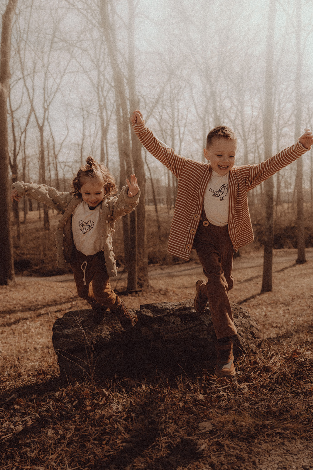 Two children balancing on a tree stump in a forest, arms raised, smiling, with trees in the background.