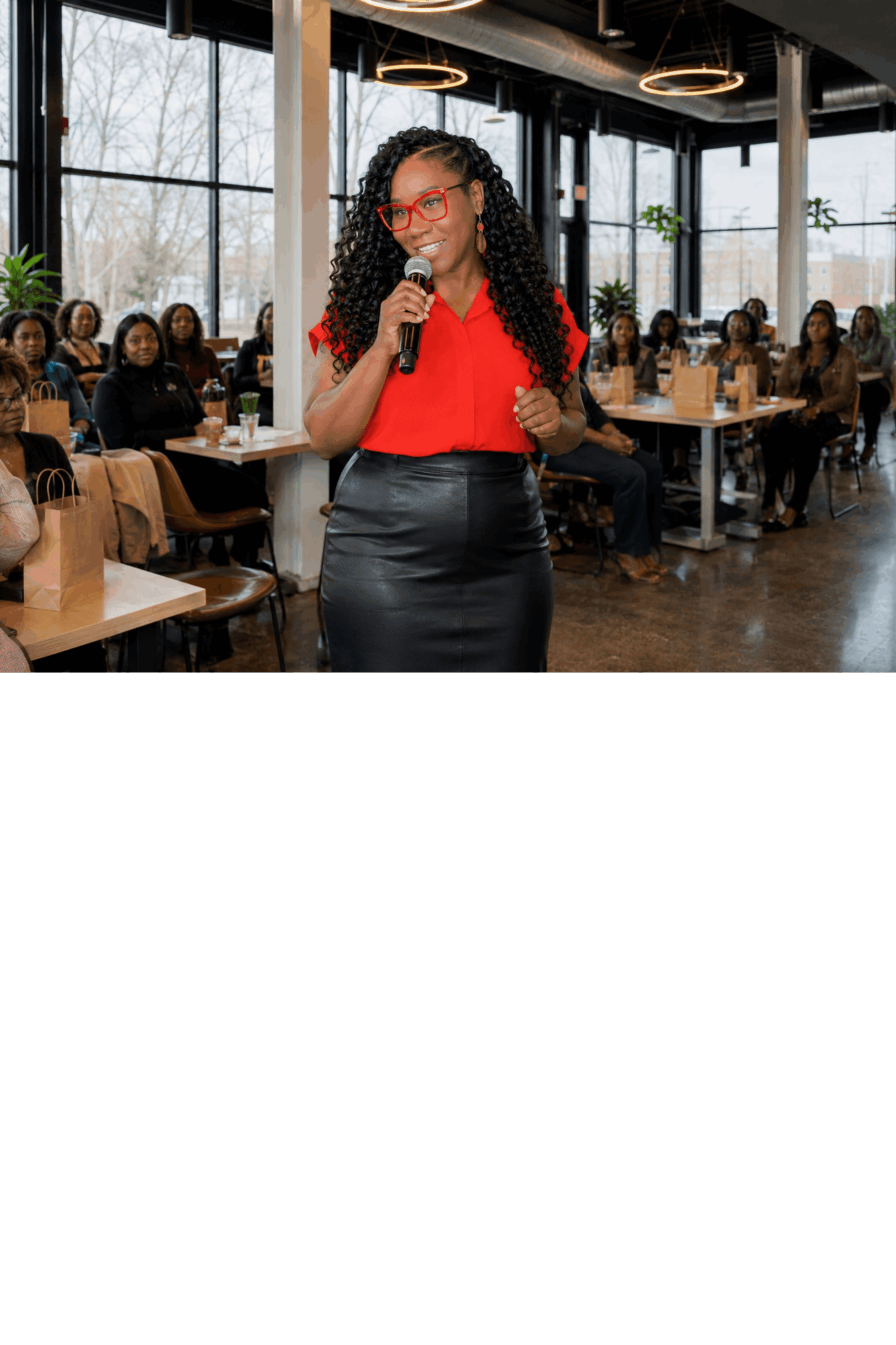 Woman with curly hair and glasses speaking into a microphone in a restaurant, audience seated at tables.