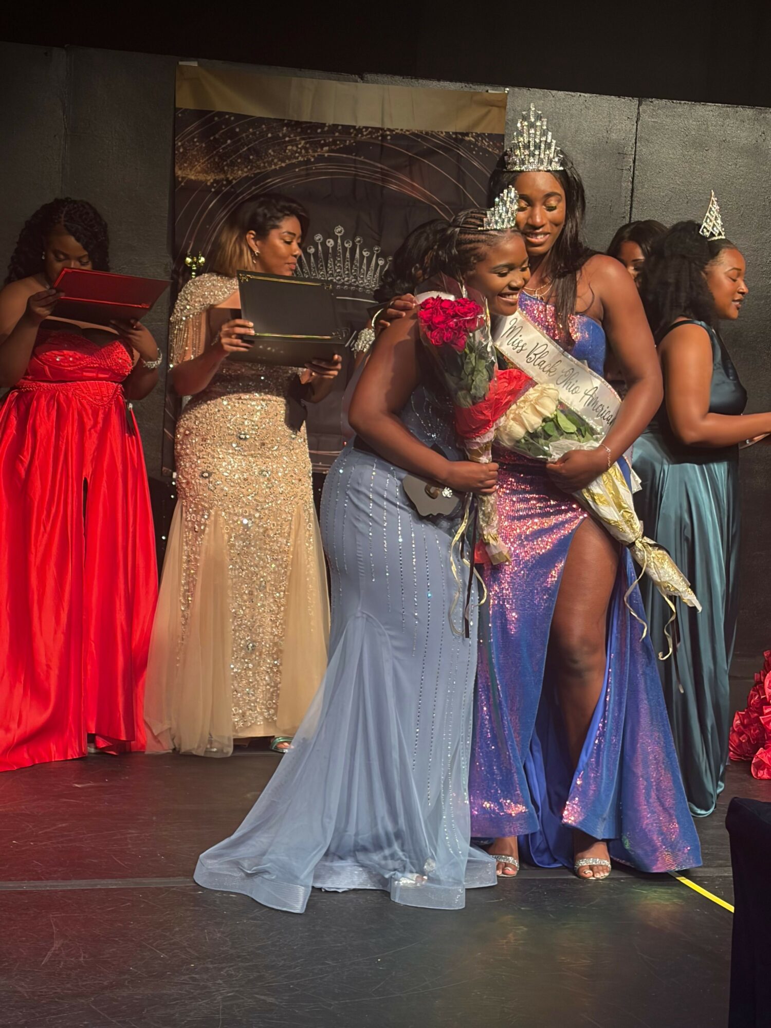 Group of women in elegant dresses, some holding bouquets, standing on stage with a dark backdrop.
