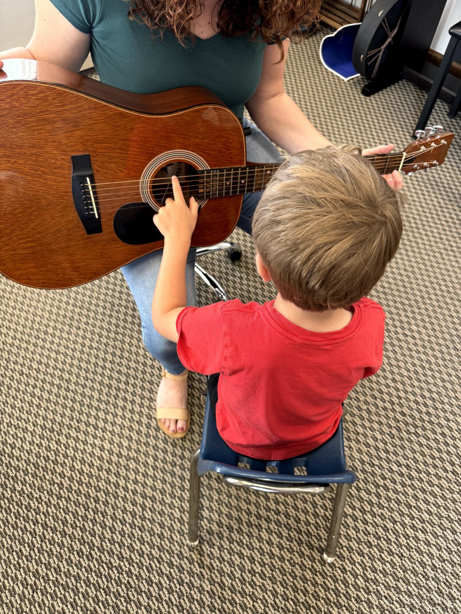 Child sitting on a chair, reaching to touch a woman playing a guitar, in a room with patterned carpet.