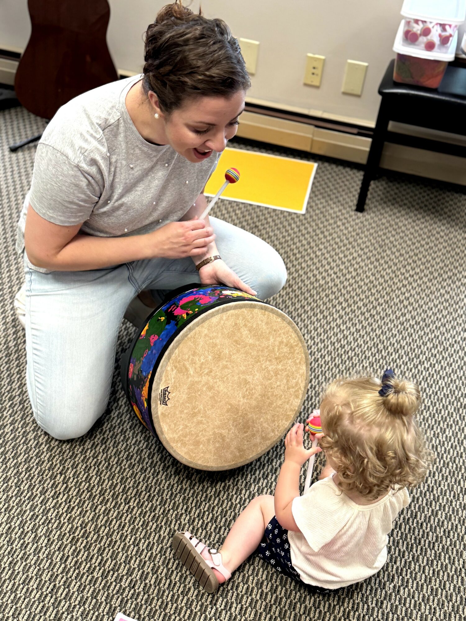 Adult and child sitting on carpeted floor, playing with a drum and a yellow book, smiling and engaging with each other.